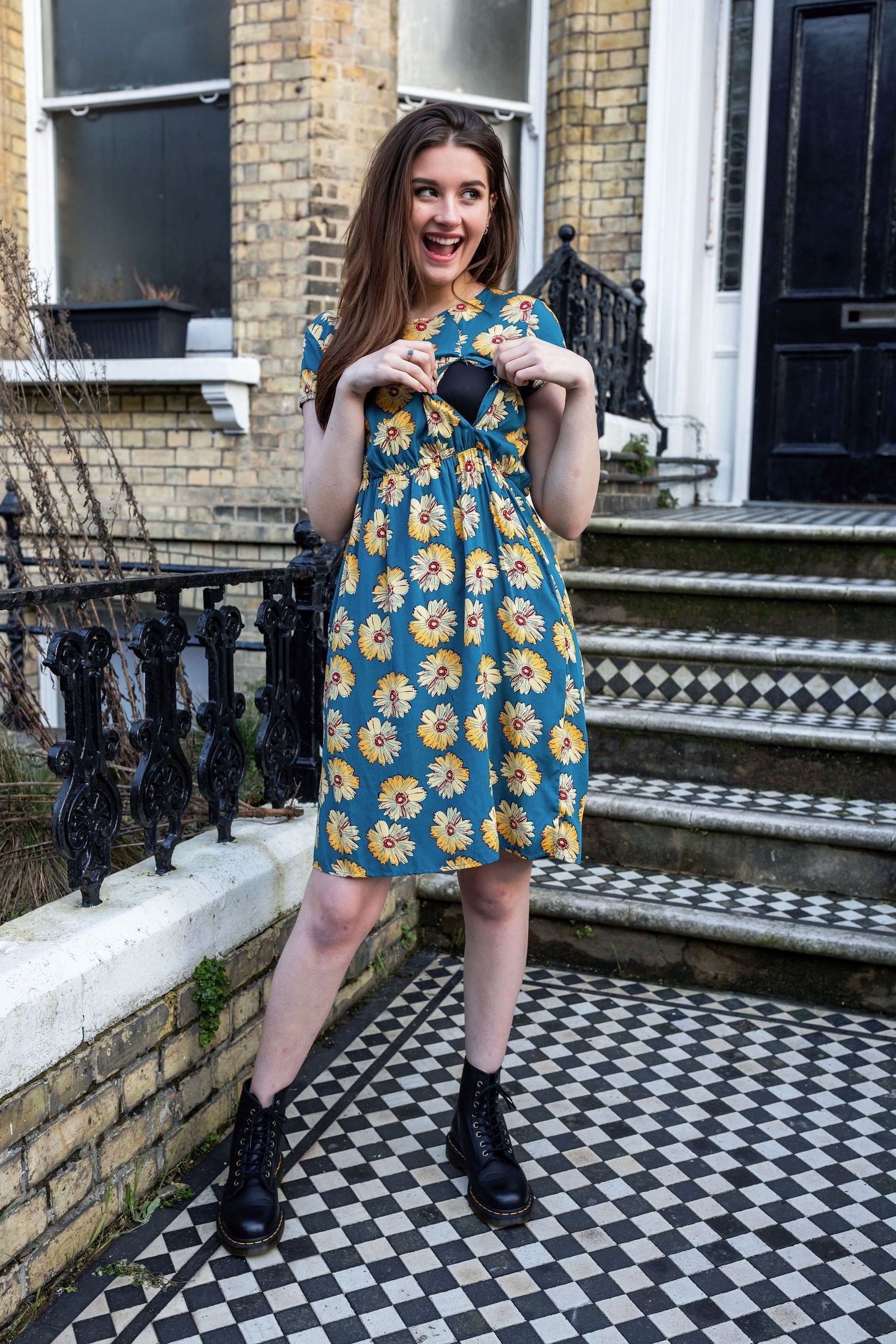 Woman in a floral breastfeeding dress standing on a checkered pavement outside a building. She is showing the breastfeeding access of the dress with her hands.