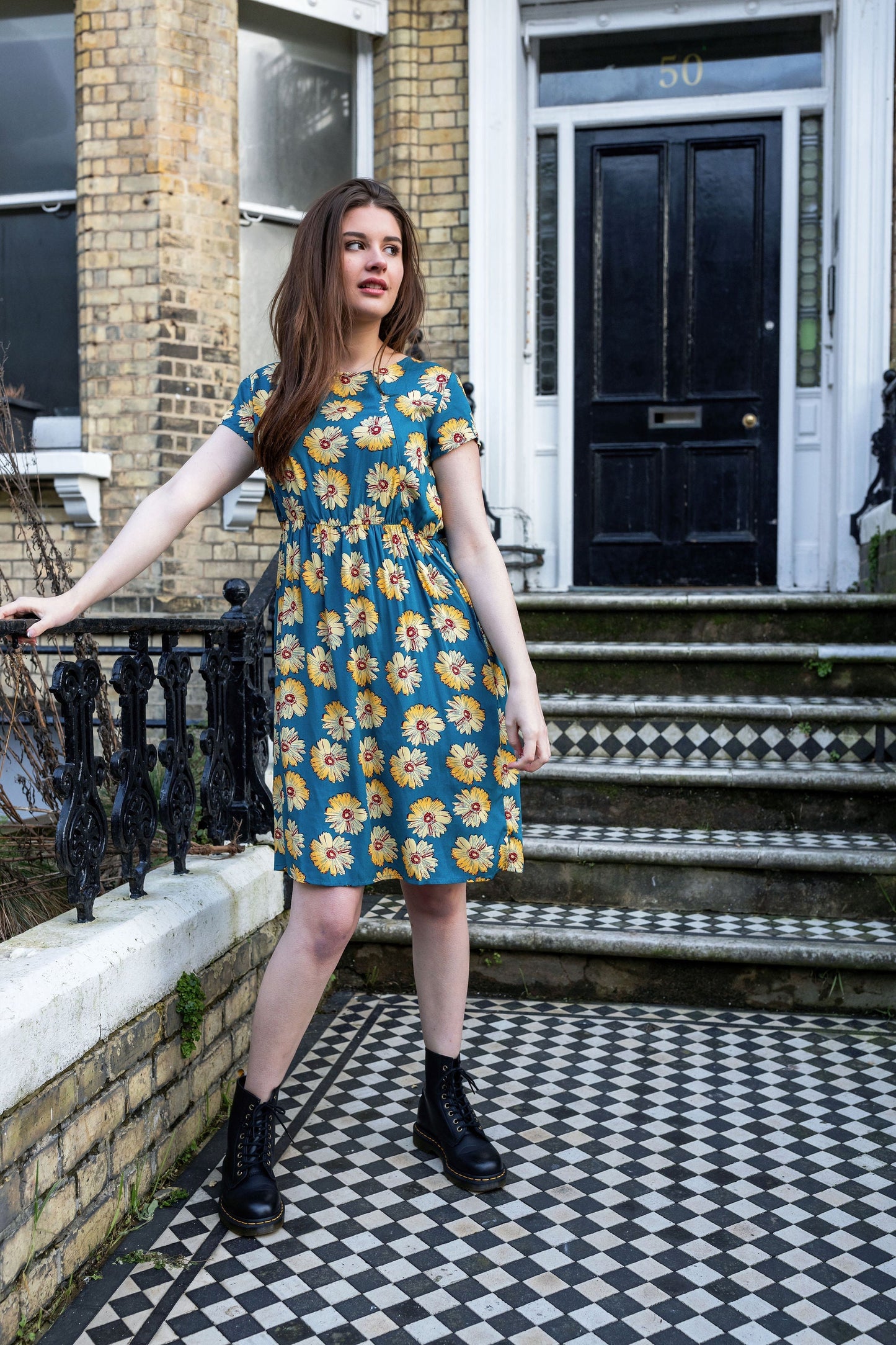A model wearing a knee-length blue breastfeeding dress with a sunflower pattern, featuring short sleeves and a Peter Pan collar, standing on a doorstep.