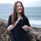 Woman in a black breastfeeding dress standing against a stone wall with ocean in the background showing breastfeeding access via a zip.