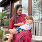 Mum with long dark hair sat on bench outside breastfeeding her toddler. She is wearing a beautiful red leopard print long midi dress. The dress has hidden zips for breastfeeding so although the mum is breastfeeding it is very discreet. The dress is a best seller by Stylish Mum which is a breastfeeding UK brand. 