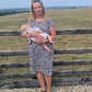 Woman breastfeeding a baby in a patterned breastfeeding dress standing near a wooden fence with a scenic background.